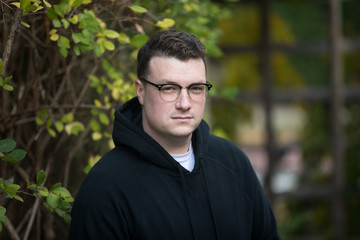 A caucasian male wearing a black hoodie sweater and glasses poses in a garden while surrounded by fall foliage on a cold afternoon.