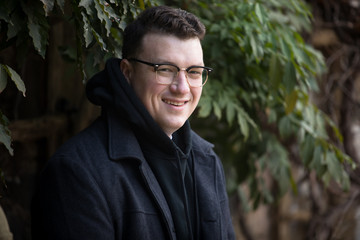 A caucasian male wearing a black hoodie sweater and glasses poses in a garden while surrounded by fall foliage on a cold afternoon.