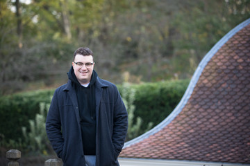 A caucasian male wearing a black hoodie sweater and glasses poses in a garden while surrounded by fall foliage on a cold afternoon.