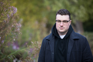 A caucasian male wearing a black hoodie sweater and glasses poses in a garden while surrounded by fall foliage on a cold afternoon.