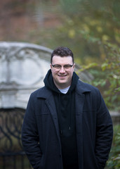A caucasian male wearing a black hoodie sweater and glasses poses in a garden while surrounded by fall foliage on a cold afternoon.