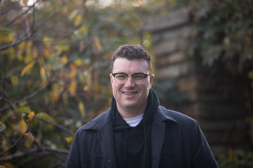 A caucasian male wearing a black hoodie sweater and glasses poses in a garden while surrounded by fall foliage on a cold afternoon.