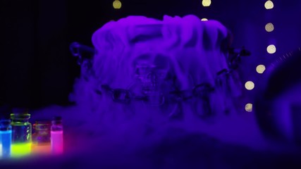 Halloween Bubbling smoking witch's cauldron with neon glowing vials and crystal ball in foreground. A hand empties poison into the pot. Witch and wizard concepts. RED Camera, Slow motion. - Powered by Adobe