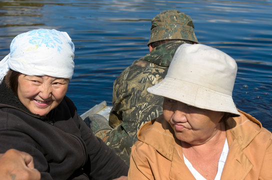 Faces joyful and sad two Yakut women sitting in a boat floating on the river with men.
