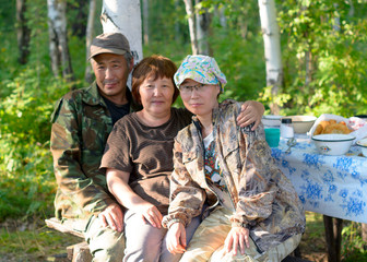 An elderly couple of rural Asian Yakuts and a young girl family pose on a bench at a table in the open air forest in the Northern taiga of Yakutia.