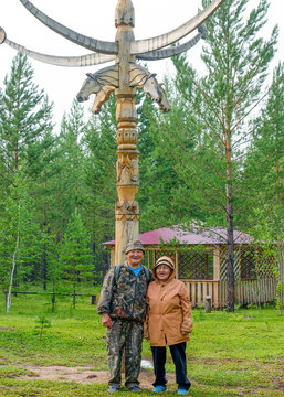 Two elderly Yakuts a man and a woman stand hugging and smiling under the national ritual pillar of Serge in the Park.