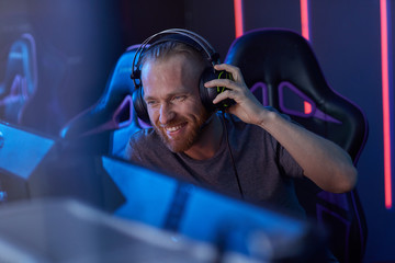 Happy bearded young gamer in headphones looking at computer monitor and laughing while sitting in...