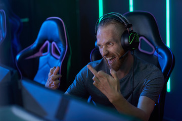 Young man in headphones looking at computer monitor and gesturing he teasing his rival while playing computer game