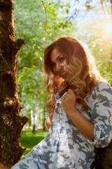 Beautirul girl with curly blonde hair near a tree in the park with sunny weather