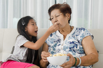 Asian senior woman eating popcorn with her grandchild while watching TV at home,