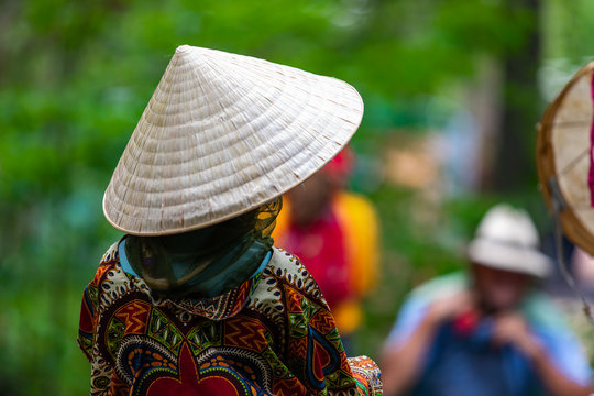 close up and selective focus on a woman wearing Asian conical hat viewed from the back, with copy space and blurry background of people and nature