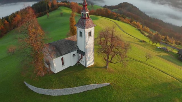 Aerial view of the hills, colorful forest in the fog and the church of Sv Tomaz. Sunrise in Slovenia in autumn