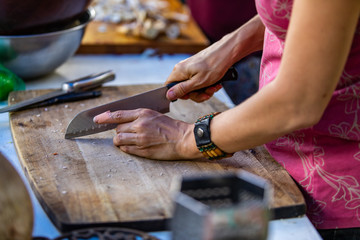 Woman responsible of a festival camp kitchen is seen from the left as she cuts vegetables on a big wooden slate of wood with a chopping knive. 