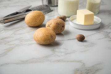 Kartoffeln, Milch, Salz, Butter, Muskatnuss als Zutaten mit Kartoffelpresse und Muskatreibe für Kartoffelpüree auf hell Marmor Hintergrund