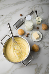 Kochtopf mit Kartoffelpüree mit Zutaten aus Kartoffeln, Milch, Salz, Butter, Muskatnuss dazu Muskatreibe und Schneebesen auf hell Marmor Hintergrund