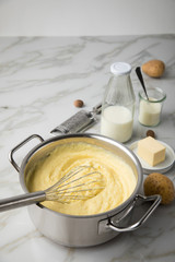 Kochtopf mit Kartoffelpüree mit Zutaten aus Kartoffeln, Milch, Salz, Butter, Muskatnuss dazu Muskatreibe und Schneebesen auf hell Marmor Hintergrund