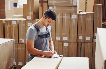 Young male worker in uniform is in the warehouse with notepad and pen