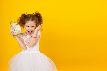 Image of little girl child standing isolated over yellow background, holding a clock alarm, looking at camera with open mouth, have a scream expression.