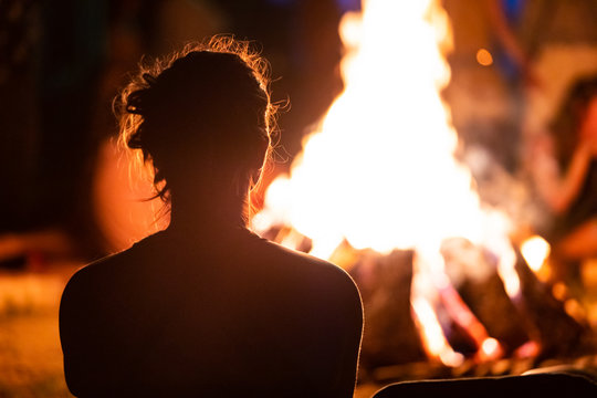 silhouette of a man with long hair sitting in the front of the fire, seen from behind during dark night campfire, blurred background