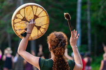 A spiritual redhead woman is viewed from behind as she raises a native drum and drumstick with...