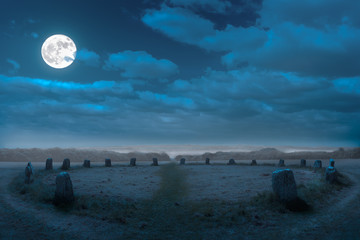 The mystical historic stone circle of Merry Maidens in Cornwall at night. It is foggy and the full moon is shining in the sky. Some clouds are in the sky.