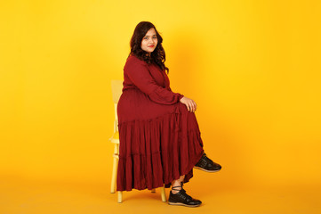Attractive south asian woman in deep red gown dress posed at studio on yellow background and sitting on chair.