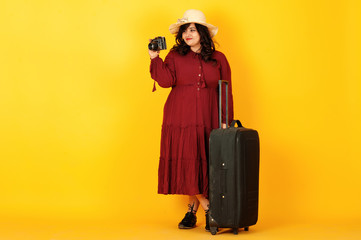 Attractive south asian traveler woman in deep red gown dress, hat posed at studio on yellow background with suitcase amd old vintage photo camera.