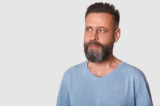 Studio Shot Of Attractive Swedish Guy With Stylish Haistyle And Beard Wearing Grey T Shirt, Looks Thoughtful, Looking Aside Thinking About Something. Copy Space For Advertisment Or Promotion Text.