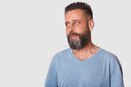 Picture of handsome bearded man standing and looking aside with serious facial expression, wears glasses and casual gray t shirt, model posing isolated over white background. Copy space for promotion.