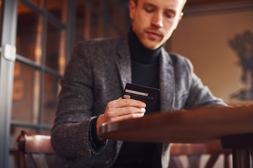 Portrait of modern young guy in formal clothes that sits in the cafe and holds credit card in hand