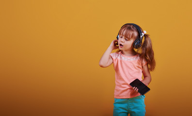 Portrait of cute little girl in headphones and with phone in the studio against yellow background