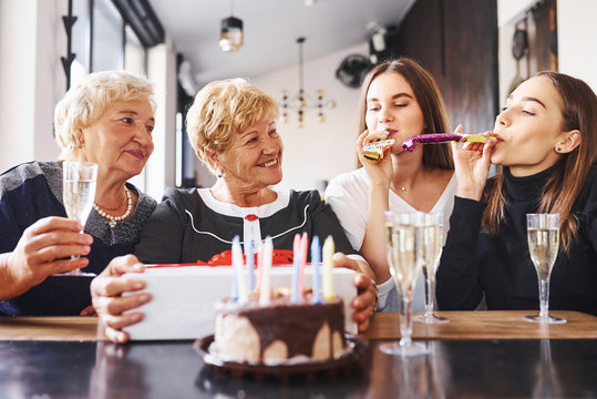 Glasses with alcohol in hands and cake on table. Senior woman with family and friends celebrating a birthday indoors