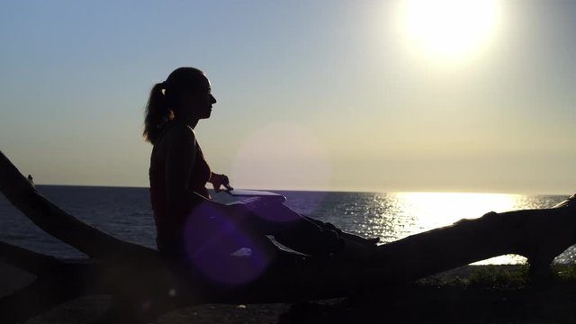 silhouette of girl opens laptop and works on it in patch of reflected light of sun at sunset on seashore.