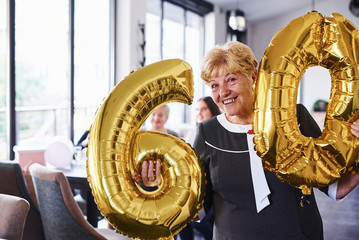 With balloons of number 60 in hands. Senior woman with family and friends celebrating a birthday indoors