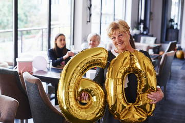 Fototapeta na wymiar With balloons of number 60 in hands. Senior woman with family and friends celebrating a birthday indoors