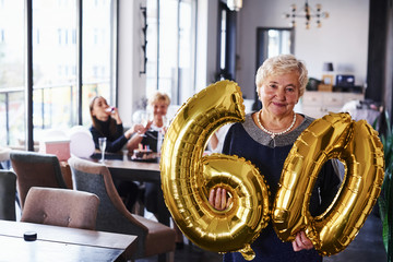 With balloons of number 60 in hands. Senior woman with family and friends celebrating a birthday indoors