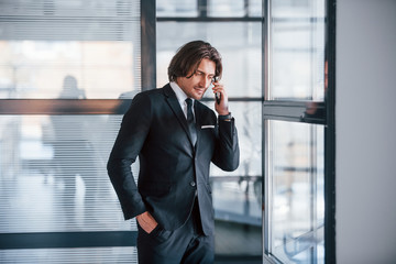 Talking by the phone in the office. Portrait of handsome young businessman in black suit and tie