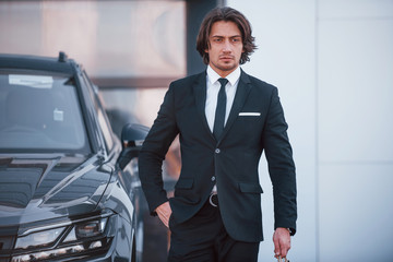 Portrait of handsome young businessman in black suit and tie outdoors near modern car