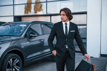 Portrait of handsome young businessman in black suit and tie outdoors near modern car and with shopping bags
