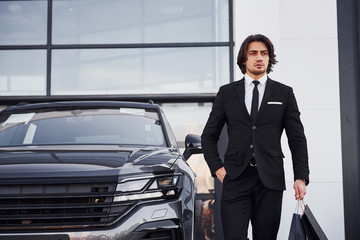 Portrait of handsome young businessman in black suit and tie outdoors near modern car and with shopping bags