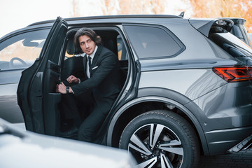 Fototapeta na wymiar Walks out from car. Young businessman in black suit and tie inside modern automobile