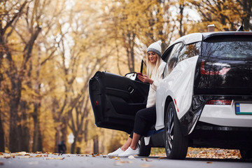 Using smartphone. Girl have autumn trip by car. Modern brand new automobile in the forest