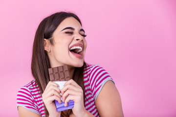 Happy young beautiful lady eating chocolate and smiling. Girl tasting sweet chocolate. Young woman with natural make up having fun and eating chocolate isolated on pink background