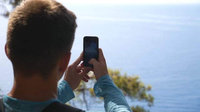 Unrecognizable man taking photo of scenic seascape on his smartphone. Young guy using phone to get beautiful pictures of sea from hill top. Male hiker enjoying travel during summer vacation. Slow mo