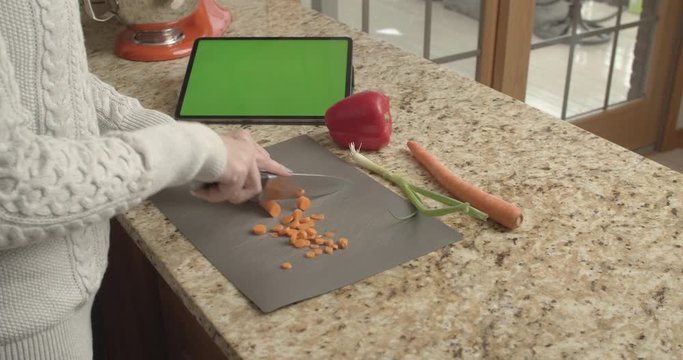 Woman chopping carrots in kitchen with a tablet with a green screen.