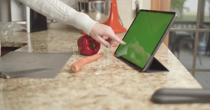 Woman in kitchen using tablet with green screen and prepping food.