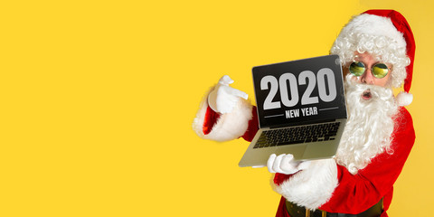 Santa Claus in sunglasses isolated on yellow studio background. Caucasian male model in traditional costume. Concept of Christmas, 2020 New Year's, winter mood. Copyspace. Showing laptop screen.