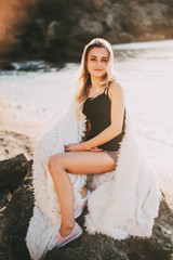 Beautiful girl with beautiful hair in shorts sitting on a rock on the background of rocks and the sea or ocean, wrapped in a fluffy light plaid