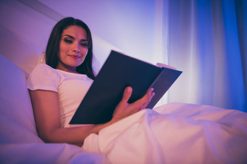 Low below angle view portrait of her she nice attractive lovely charming sweet calm peaceful girl lying in bed reading novel story spending night in illuminated room house hotel flat apartment