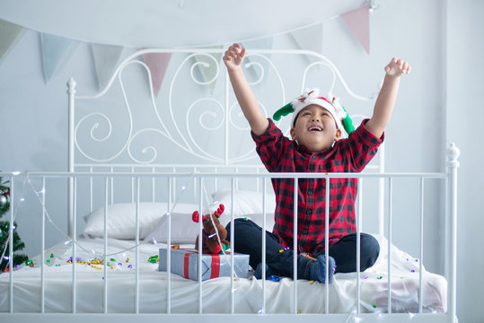 Asian Boy Wearing A Party Hat Sat On The Bed And  Raise Hand Up Feels Happiness, It's  Christmas Time